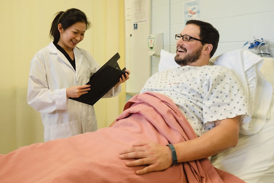 A nurse holds a clipboard and stands beside a patient in a hospital bed